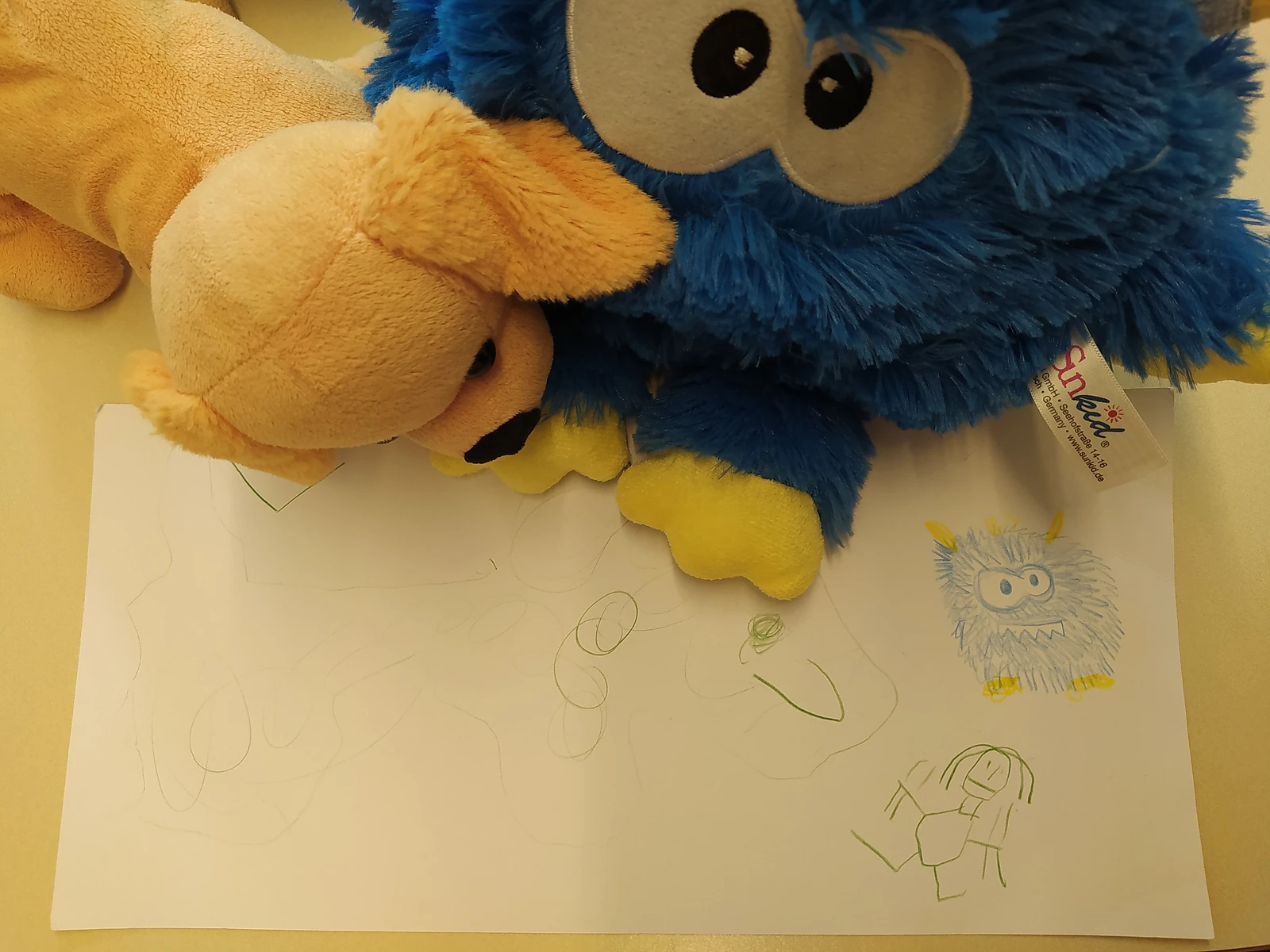 ein blaues, rundes Kuscheltier mit großen Augen sitzt mit einem Stofftier Hund auf einem Tisch. Vor ihnen liegt ein Blatt Papier auf dem ein Kind gemalt hat.